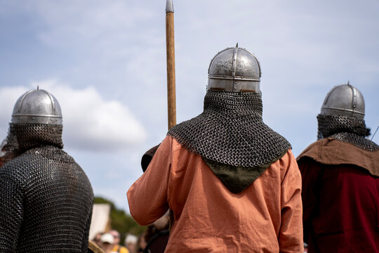 Medieval warriors in steel helmets with chainmail aventails standing in formation holding a spear outdoors under daylight.