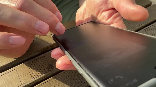 Smartphone user tapping touchscreen, quick reply online. Adult man holding modern mobile phone outdoors at wooden table background. Fast texting in mobile chat application during day time
