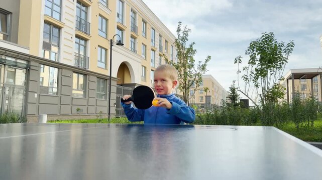 A focused young boy holds an orange ball against his paddle. He prepares to serve table tennis at an outdoor court near modern buildings. His eager expression shows pure delight and determination.