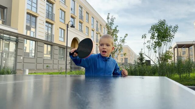 A focused young boy holding a racket watches an orange ping pong ball fly. He plays table tennis at an outdoor court near modern buildings. His eager look shows pure determination and joy.
