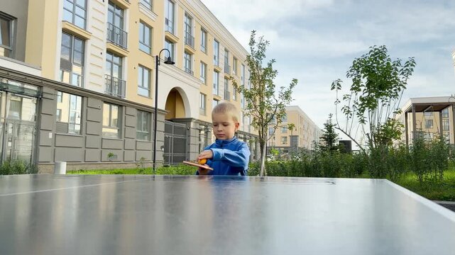 A focused little boy prepares to serve in table tennis with calm determination. He stands at an outdoor table in a modern residential courtyard, filled with eager joy.
