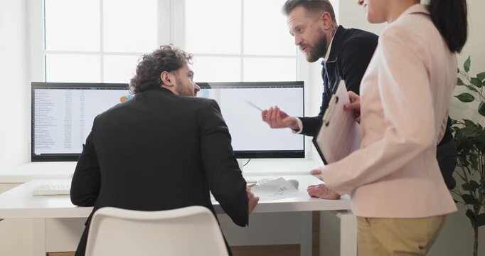 Team meeting in office reviews computer data analytics. A seated man consults colleagues and a woman, reviewing reports on monitors and aligning plans. Good teamwork and planning concept.