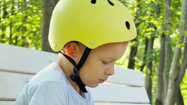 A young boy wearing a yellow helmet rests quietly on a white bench. He looks down with calm focus amid lush green trees.