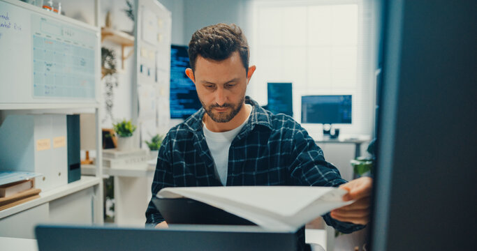 Young Caucasian male programmer writes code using multiple screens in home office workspace. AI development, cybersecurity, network database system, software engineering, remote tech startup concept.