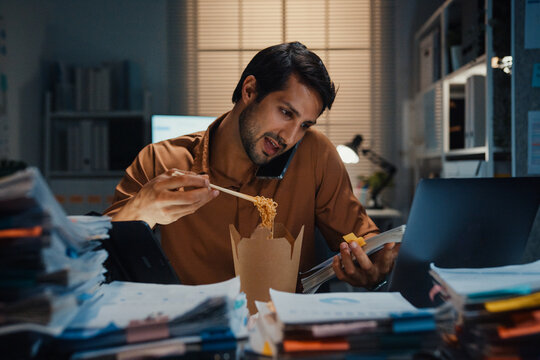Young Latin businessman taking urgent phone call while reading accounting document on laptop at desk in dark night office. Multitasking overtime, overworked deadline, tired paperwork rush concept.