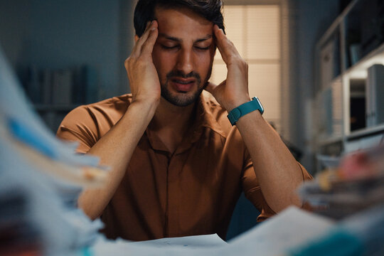Stressed young Latin businessman having severe headache and migraine at desk in night office. Debt crisis, corporate burnout, late overtime workload, mental health anxiety, business failure concept.