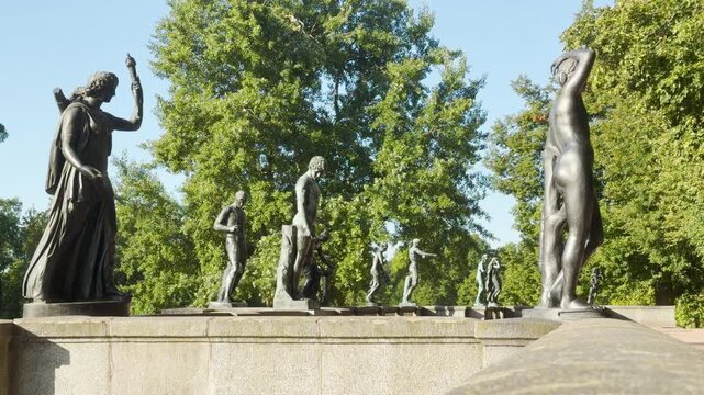 Bronze statues stand gracefully along a stone terrace in Catherine Park, Saint Petersburg. Lush green trees surround the historic site. The noble sculptures inspire quiet awe.