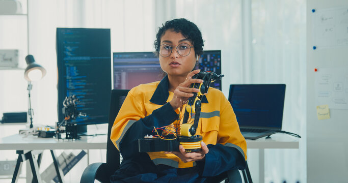 Young African American female robotics engineer discussing mechanical part on video call and looking at camera in laboratory. Remote work, virtual training, online meeting, future technology concept.