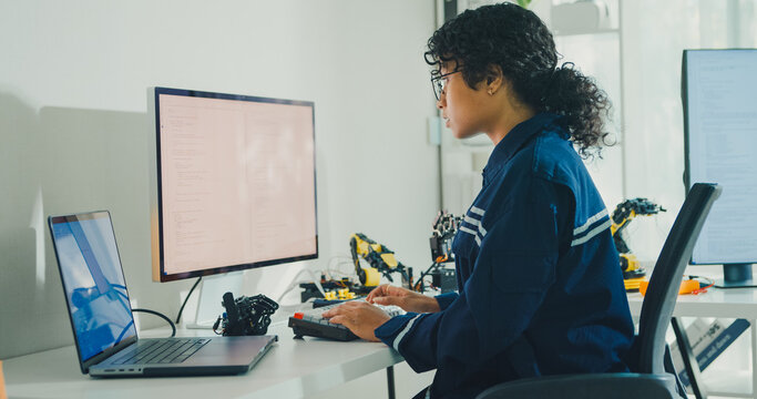 Young African American female robotics engineer coding AI software for robotic arm in laboratory. Embedded system, firmware testing, machine learning, industrial automation, future technology concept.