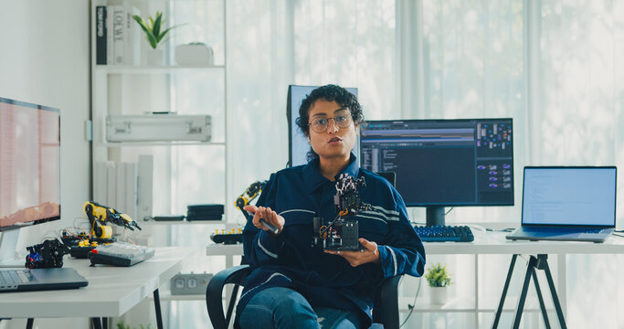 Young African American female robotics engineer discussing mechanical part on video call and looking at camera in laboratory. Remote work, virtual training, online meeting, future technology concept.