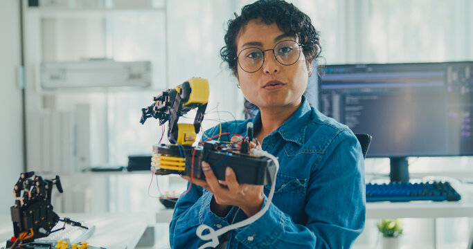 Young African American female robotics engineer discussing mechanical part on video call and looking at camera in laboratory. Remote work, virtual training, online meeting, future technology concept.