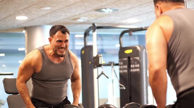 Athletic man with intense facial expression doing dumbbell lateral raises in front of a gym mirror during shoulder strength training