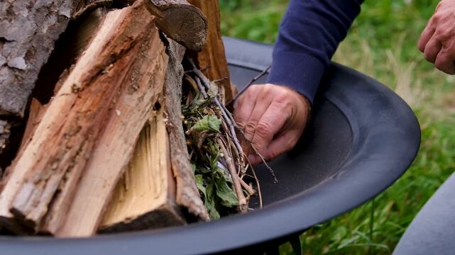 A man uses a burning match to light paper and dry kindling inside a metal fire pit beside a house, starting flames for outdoor barbecue preparation. Close up.