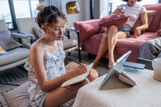 Woman Writing in Notebook While Friends Relax on Sofa