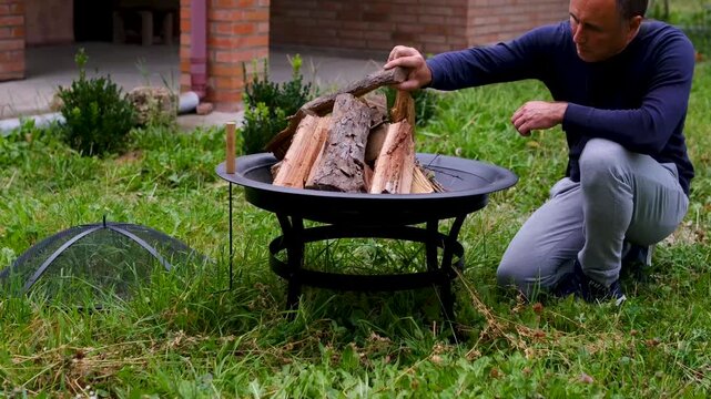 A man places large split logs inside a metal fire pit on green grass, carefully arranging firewood before lighting an outdoor fire near a country house. Close up.