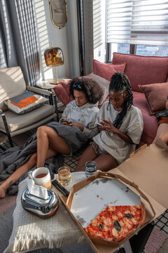 Two women relax at home with pizza and mobile phones