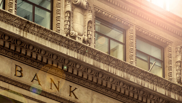 Historical bank building exterior with ornate stone architectural detailing under bright sunlight, captured in an upward perspective shot b 26