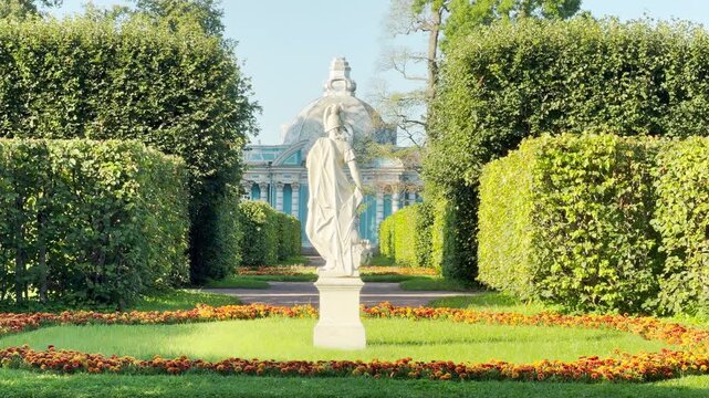 A marble statue stands gracefully among trimmed hedges in Catherine Park, Saint Petersburg. The Grotto pavilion rises peacefully in the background. This elegant garden brings a joyful mood.