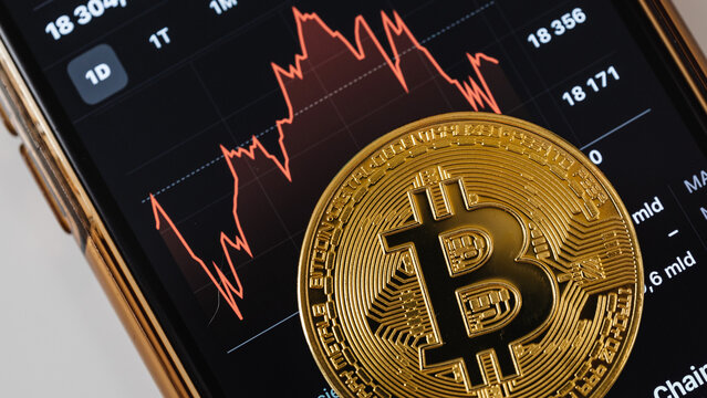 Cryptocurrency market analysis: A physical gold Bitcoin token placed on a smartphone screen displaying a fluctuating digital trading chart