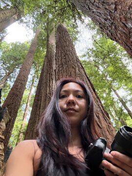 Woman Standing Under Tall Trees in a Forest Holding a Camera