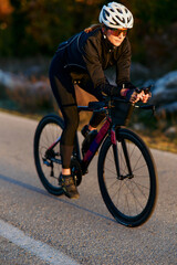 Fototapeta premium Female Cyclist On Road Bike Training At Sunset Wearing Helmet And Sport Sunglasses