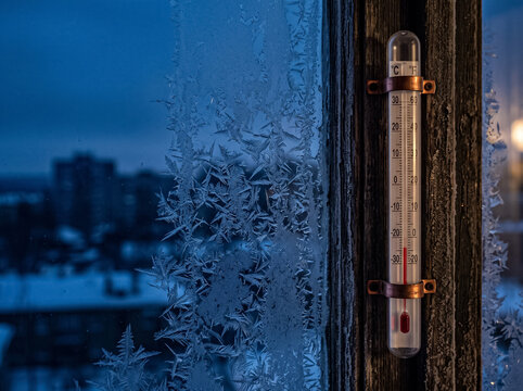 thermometer behind frozen window shows extreme winter cold background of snowy city buildings at twilight atmosphere of freezing weather on street
