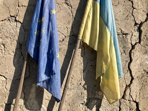worn and damaged flags of european union and ukraine are fixed on wooden poles near a cracked dry clay wall under bright sun showing resilience of allies during hard times