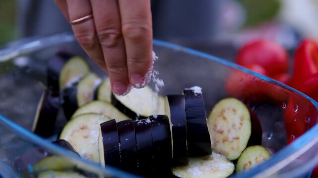 Female hands prepare eggplants for outdoor grilling by slicing, salting, rinsing and squeezing out excess moisture in a glass bowl before cooking on barbecue. Close up.