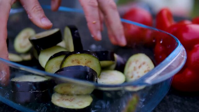 Female hands prepare eggplants for outdoor grilling by slicing, salting, rinsing and squeezing out excess moisture in a glass bowl before cooking on barbecue. Close up.