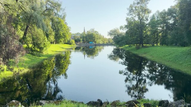 A tranquil canal reflects lush green trees and a distant spire in Catherine Park, Saint Petersburg. The peaceful scenery brings a sense of calm and wonder.