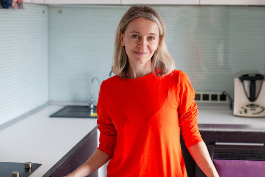 Smiling mature woman in red shirt standing in modern kitchen at home. Happy middle aged housewife cooking meal in apartment. Domestic life, housework and cozy interior concept.