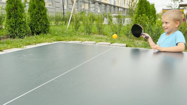 A smiling young boy holds his paddle with joyful excitement to play table tennis. He stands at an outdoor table in a green courtyard.