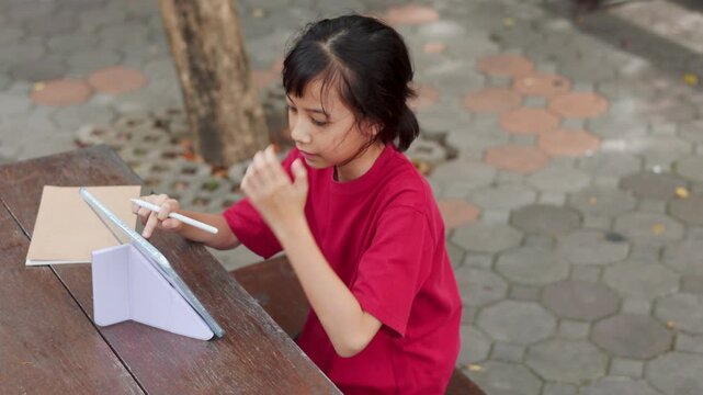 Asian kid using stylus pen on a digital tablet for e-learning at outdoor school table. Selective focus shows the young student engaging with technology for modern education and digital study concepts.