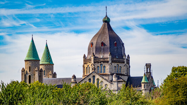 Two towers with green domes and grey cupola of central nave of St. Lambert's Church stand out among green foliage of trees, sunny day with blue skies in Maastricht, South Limburg, Netherlands