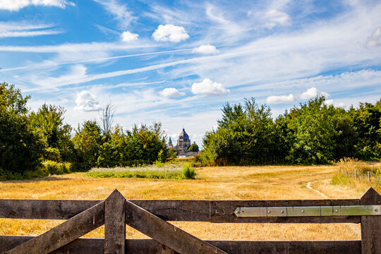 Plain with dry grass surrounded by leafy green trees in Fronten Park with St. Lambert Church in background, sunny day with blue sky and white clouds in Maastricht, South Limburg, Netherlands