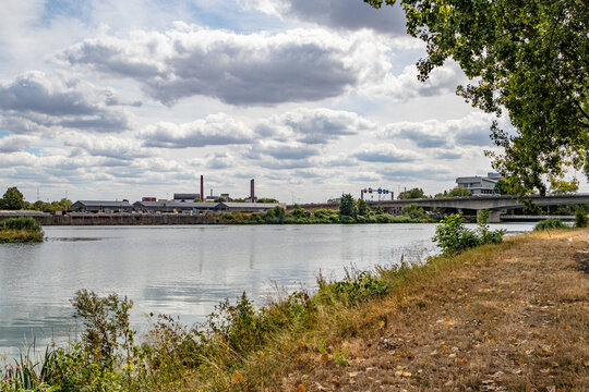 Meuse River with industrial land and vehicular bridge in urban view in background, riverbank with dry grass, cloudy summer day against sky with abundant clouds, Maastricht, South Limburg, Netherlands