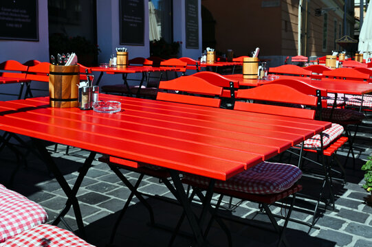 restaurant terrace or patio with bright red wood tables and chairs. glass ashtray, salt and pepper shakers. cutlery in wood drums. tasteful presentation. bright summer light. european city scene.  