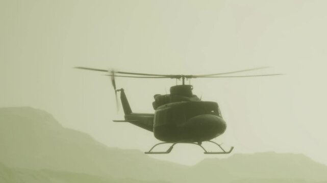 A helicopter is flying above a mountain range on a hazy afternoon. The rotors spin fast as it moves through the sky. The landscape below shows peaks partially obscured by haze.