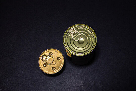 Two different sized tin cans with tin rings for opening stand on a thick kitchen table.