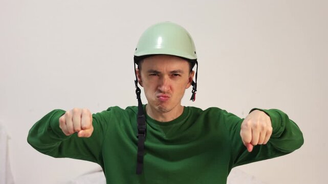 Funny man wearing a green helmet and pullover pretending to ride a motorbike or drive a cycle, making comical faces and winking while looking directly into the camera, isolated on a white background