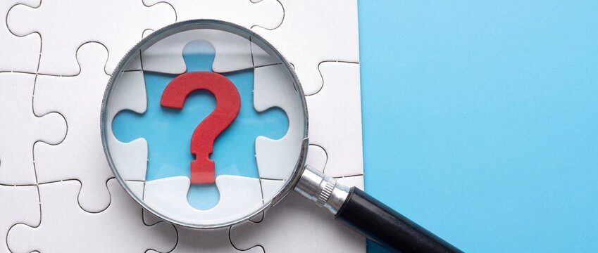 A red question mark symbol viewed through a magnifying glass on top of white puzzle pieces with symbol blue background rests white jigsaw close up investigation with information