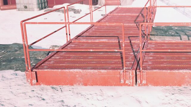 A red metal walkway sits on a snowy landscape. It is placed close to large industrial buildings. The ground shows patches of snow and rock in the outdoor setting.