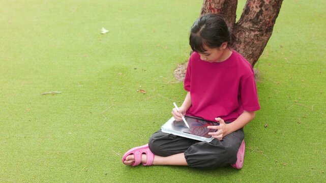 Asian kid sitting under a tree on the grass, drawing digital art on a tablet. Selective focus highlights the creative elementary student enjoying a relaxing outdoor hobby and personal time.