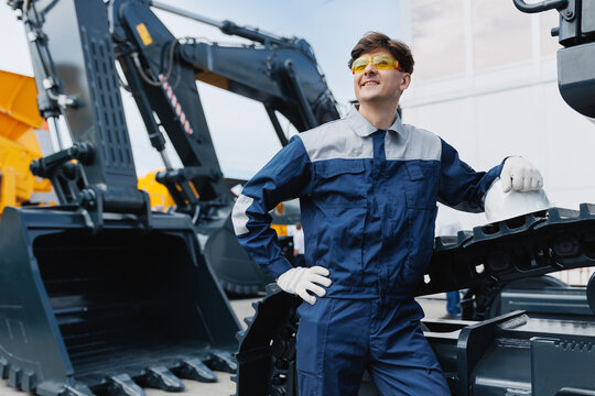 Happy Caucasian young male driver of crawler excavator with hard hat standing by mining equipment
