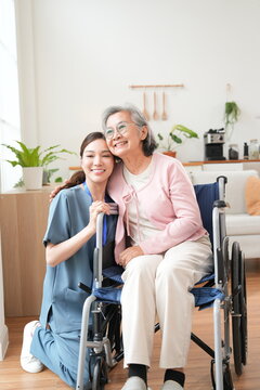 An elderly female patient with Alzheimer's disease is in a wheelchair, with an Asian nurse sitting beside her, holding her hand, offering encouragement, and pushing the wheelchair, caring for the elde