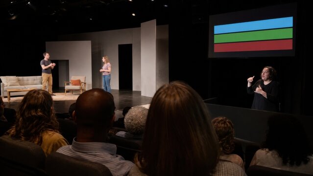 Deaf audience members watch a small theater performance while a sign-language interpreter works in a lit side area and a caption screen shows only colored timing bars