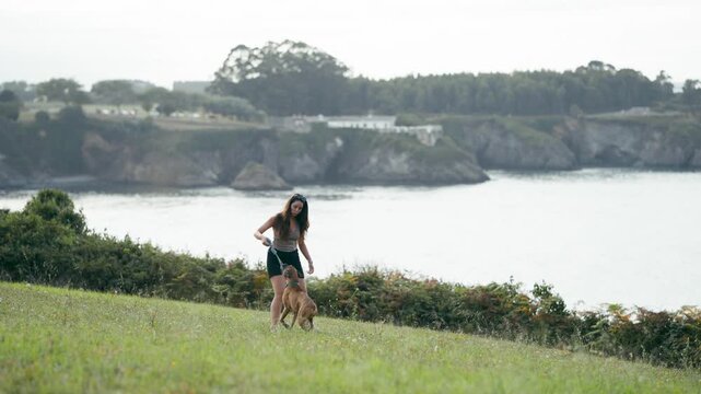Happy young woman walking and playing with her boxer dog on a leash in a green meadow with a beautiful coastline view in the background