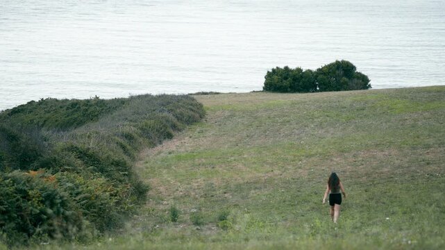 Solitary young woman with long dark hair walking away through a grassy meadow. A scenic coastal landscape with green hills and the ocean