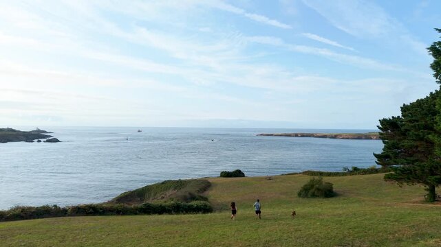 Young couple with a small dog enjoying a summer day running together in a green field overlooking the ocean, embracing an active lifestyle