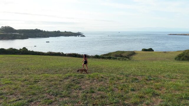 Beautiful young woman enjoying a relaxing walk with her small dog on a grassy hill overlooking a scenic bay with boats on a sunny day